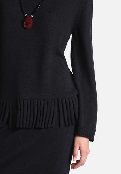Pull en maille noir avec ourlet à franges porté avec une jupe assortie et un collier pendentif en pierre rouge sur un cordon noir.