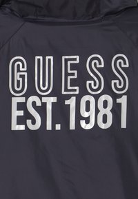 Chaqueta navy con texto en plata destacado "GUESS" y "EST. 1981" en la parte posterior. Textura suave con un material ligero y resistente al agua.