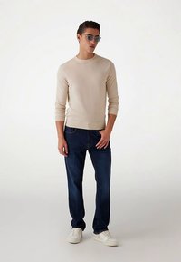 Bēša garroku džemperis, tumši zili džinsi, balti sporta apavi un apaļas saulesbrilles. Gluda tekstūra, piegulošs dizains ar ribām aprocēs.