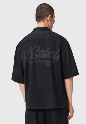 Mężczyzna w czarnej koszulce z krótkim rękawem z napisem "AllSaints enjoying the journey since '94" na plecach, stojący na szarym tle.