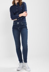 Žena stojí s překříženýma rukama, oblečená v námořnické košili na knoflíky, tmavomodrých skinny jeans a bílých vysokých teniskách.