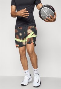 Atleta masculino vestido con camiseta negra, pantalones cortos con un patrón, zapatos y calcetines blancos de Nike, sosteniendo un balón de baloncesto negro y gris con el texto "Jordan".