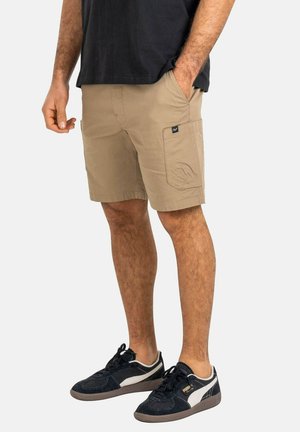 Mann trägt beige Cargo-Shorts mit Taschen, schwarze Puma-Sneaker und ein schwarzes T-Shirt, steht mit einer Hand in der Tasche vor einem schlichten Hintergrund.