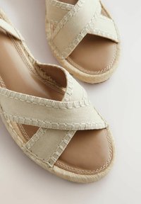 Béžové espadrilky s prekríženými plátennými popruhmi a mäkkou koženou stielkou. Obsahujú pletenú jutu na podrážke pre zvýšenie textúry a pohodlia.
