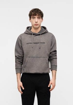 Mladi muškarac u sivoj Alpha Industries dukserici s prednjim džepom i crnim hlačama, stoji na jednostavnoj svijetloj pozadini.