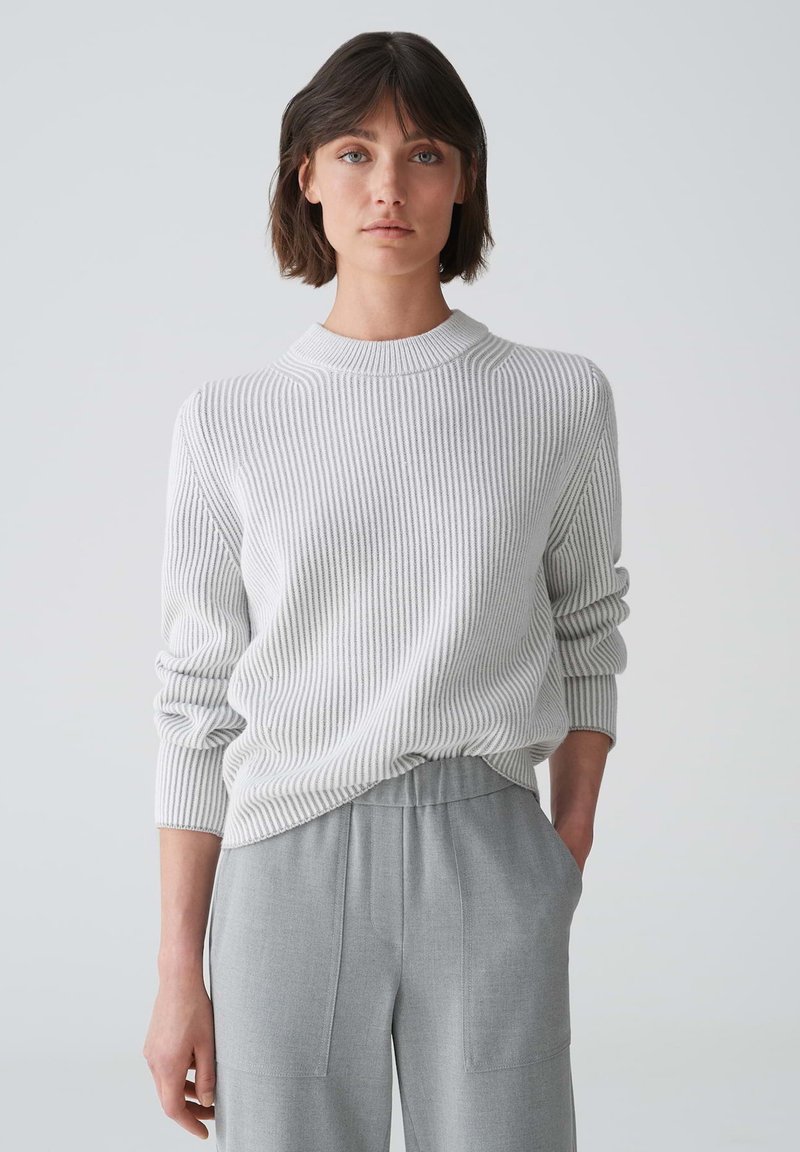 Hellgrauer, gerippter Strickpullover mit hohem Kragen und langen Ärmeln, kombiniert mit grauen, weiten Hosen mit Seitentaschen.