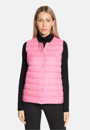 Frau, die eine ärmellose, rosa gesteppte Weste mit Knöpfen über einem schwarzen langärmligen Oberteil und schwarze Hosen trägt, vor einem einfarbigen Hintergrund stehend.