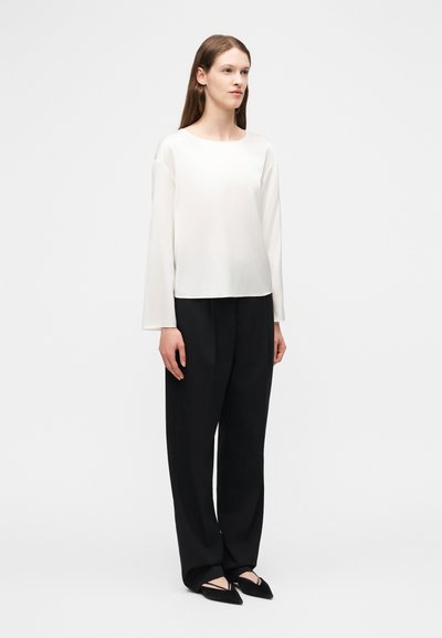 Γυναίκα που στέκεται φορώντας άσπρο πουκάμισο μακρυμάνικο, φαρδιά μαύρη παντελόνια και μαύρα μυτερά παπούτσια, σε ένα απλό λευκό φόντο.