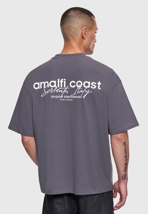 Bărbat cu păr scurt purtând un tricou gri larg cu inscripția „amalfi coast Sorrento, Italy dropsize international”, văzut din spate.