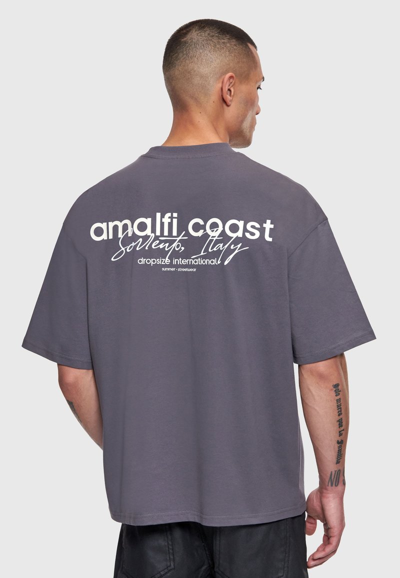 Homme aux cheveux courts portant un T-shirt gris ample avec l'inscription « amalfi coast Sorrento, Italy dropsize international », vu de dos.