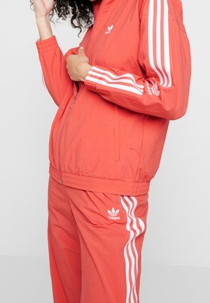 Person trägt korallenfarbenen Adidas-Trainingsanzug mit weißem Drei-Streifen-Design an Ärmeln und Hose, teilweise gezippte Jacke, Hand nahe am Gesicht.