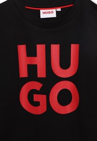 Schwarzes T-Shirt mit großen, auffälligen roten Buchstaben, die "HUGO" schreiben. Mit rundem Ausschnitt und weichem, glattem Stoff. Etikett am Ausschnitt sichtbar.