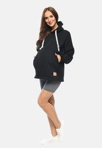 Felpa nera over size con cappuccio e cordini grigi, tasca frontale e etichetta con logo; abbinata a pantaloni corti grigi e sneakers bianche.