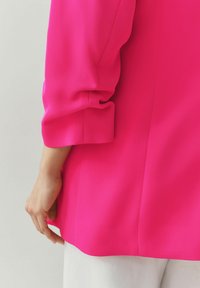 Fuchsiová blúzka z hladkej, textúrovanej tkaniny; má vyhrnutý manžet a štíhly strih, nosená cez biele nohavice.