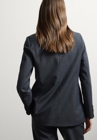 Donna con capelli castano chiaro lisci, che indossa un completo grigio scuro, in piedi con le mani in tasca su uno sfondo chiaro e semplice.