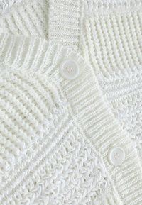Cardigan en tricot blanc avec des textures variées, notamment des motifs côtelés et en tricot ajouré, et un grand bouton blanc en détail.
