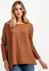 Brązowy, oversize sweter z szerokim dekoltem, długimi rękawami i ryżowym splotem. Prosty krój z bocznymi rozcięciami; noszony z niebieskimi dżinsami.