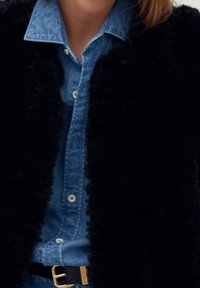 Zwarte neplederen buitenkleding over een blauwe spijkerblouse, met een klassieke kraag en knoopsluiting. Een zwarte riem met een gouden gesp maakt de look compleet.