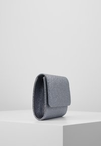 Silverglittrande handväska med vikbar klaff, rektangulär form och slät yta, som vilar på en vit pelare.