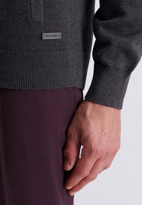 Grauer Strickpullover mit gerippten Bündchen und Saum. Verfügt über ein kleines schwarzes Logoeetikett an der Seite, kombiniert mit dunkelvioletten Hosen.