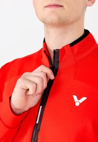 Punainen vetoketjullinen takki, jossa on korkea kaulus ja erottuva musta vetoketju. Rinnassa on valkoinen logo ja kangas on sileä ja teksturoitu.