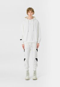 Sudadera blanca con cordones, combinada con pantalones de chándal blancos con patrones negros y detalles desgastados, llevada con botas de combate blancas.