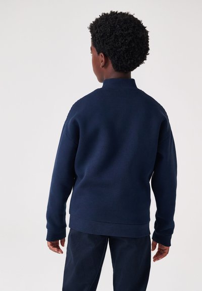 Tumši zila džemperis ar augstu apkakli, ribotu aproci un apmali. Gluda tekstūra, brīvs piegriezums, bez redzamiem logotipiem vai rakstiem.