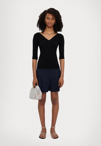 Top en mailles noires avec un décolleté en V, shorts navy, sac à main texturé argenté et sandales plates avec des brides de cheville, sur un fond neutre.