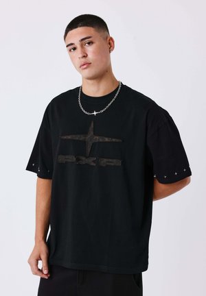 Jeune homme aux cheveux courts porte un t-shirt noir oversize avec une croix subtile et l’inscription « PXI », collier chaîne argenté, debout devant un fond uni.