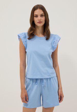 Woman wearing a light blue sleeveless top with eyelet ruffle sleeves and matching drawstring shorts, standing against a plain background.