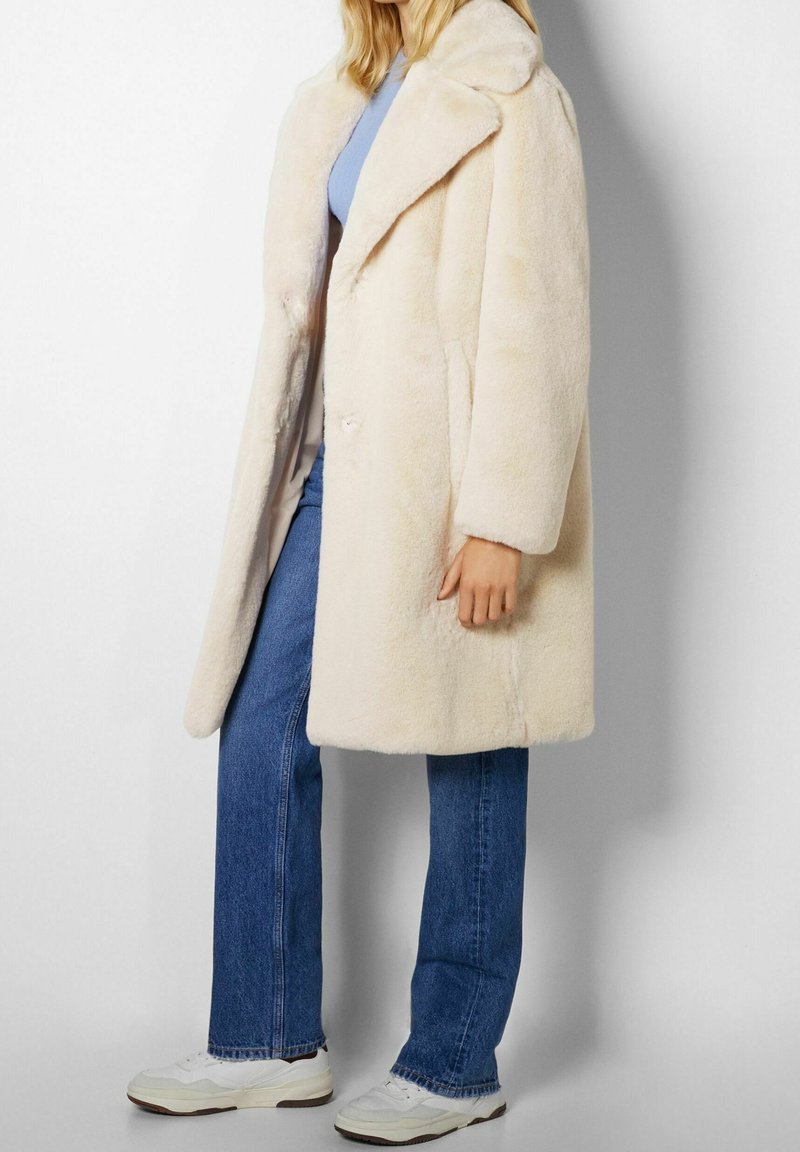 Femme portant un manteau en fausse fourrure crème longueur genou, jean bleu, baskets blanches et un haut bleu clair, debout devant un fond uni.