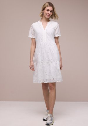 Nainen pukeutuneena valkoiseen polvipituiseen silkkiseen mekkoon, jossa on lyhyet hihat, sekä hopeisiin tennareihin, seisoo yksiväristä beigeä taustaa vasten.