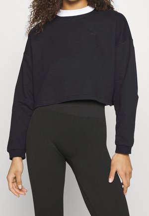 Kvinde iført en sort cropped sweatshirt over en hvid skjorte med højtaljede sorte ribbestrikkede leggings, stående mod en ensfarvet baggrund.