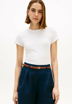 Junge Frau mit glatten braunen Haaren, die kleine goldene Creolen, ein weißes kurzärmliges T-Shirt, marineblaue hochtaillierte Hosen und einen braunen Gürtel trägt.