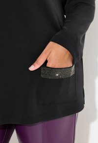 Túnica negra de manga larga con un bolsillo texturizado que presenta adornos de pedrería. Ajustada en la cintura, combinada con pantalones morados.