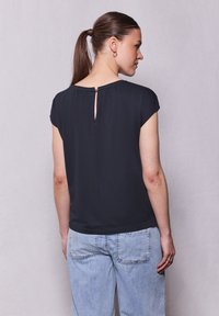 Blusa negra de manga corta con cierre en la parte posterior en forma de ojal, combinada con jeans de color azul claro. Tejido suave con un corte relajado y caída ligera.