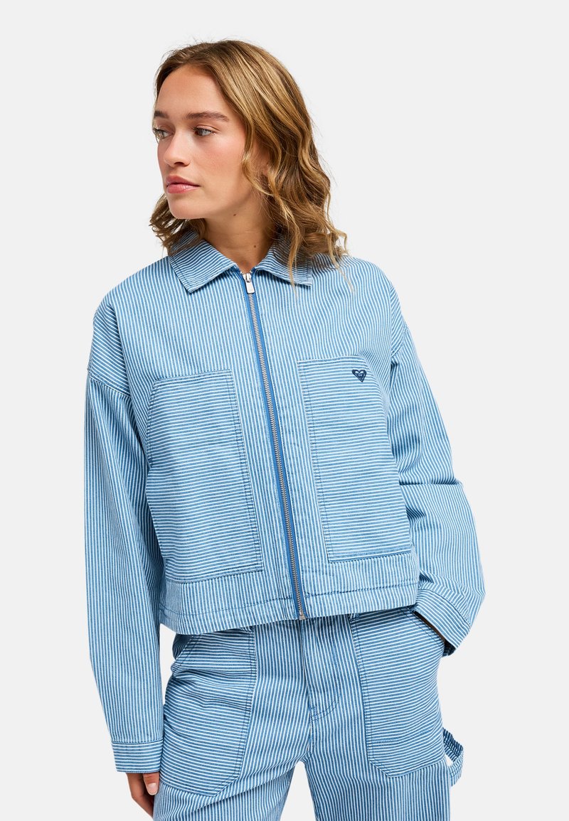 Νεαρή γυναίκα φοράει μπλε και λευκό κάθετο ριγέ μπουφάν με φερμουάρ και ταιριαστό παντελόνι, κοιτάζει στο πλάι με τα χέρια στις τσέπες.