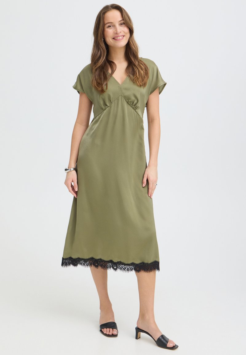 Femme souriante, portant une robe midi vert olive avec des bordures en dentelle noire et des sandales noires à talons, debout devant un fond clair uni.