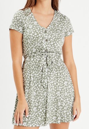 Woman wearing short-sleeve light green dress with small white floral print, front buttons, and tied waist belt.