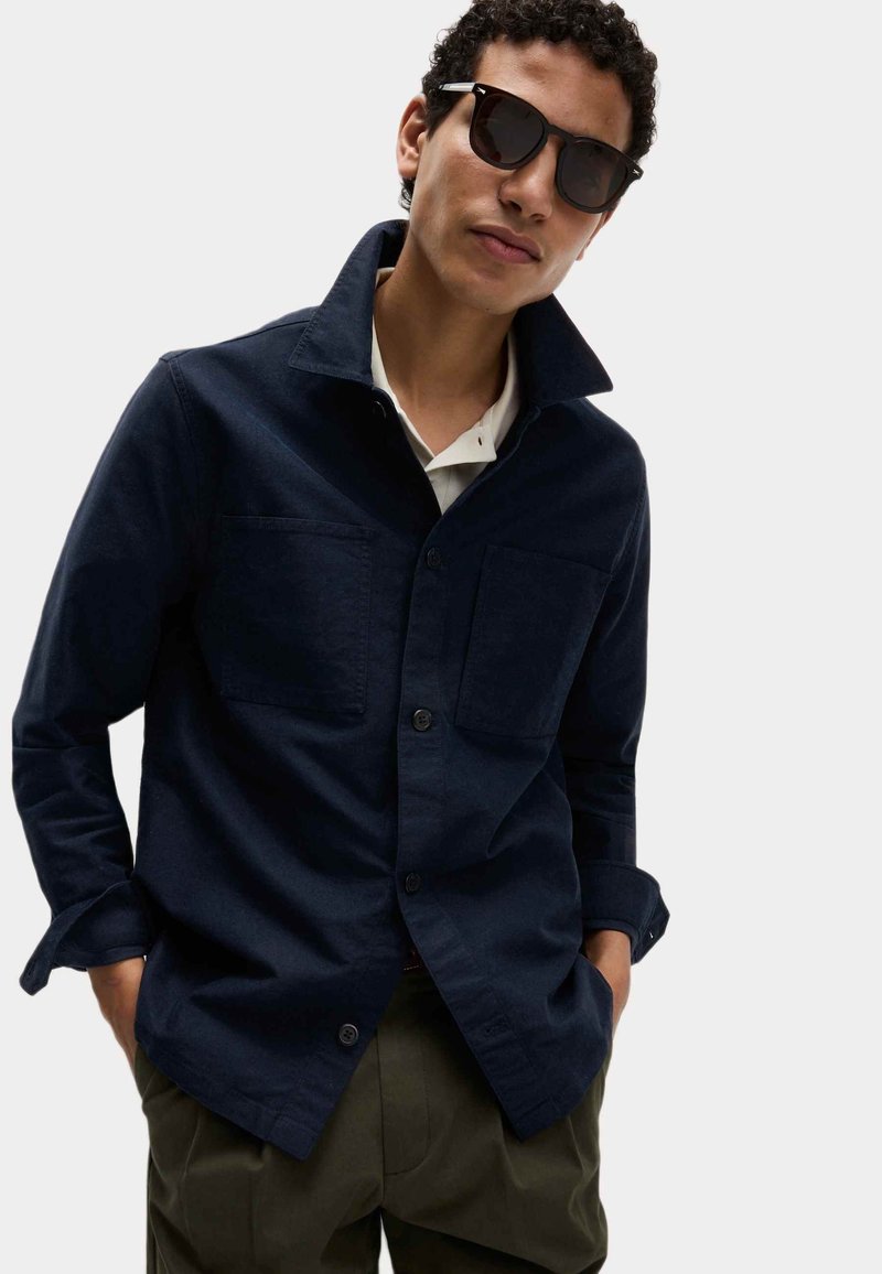 Granatowa koszula zapinana na guziki z dwiema przednimi kieszeniami, dopasowanym kołnierzykiem, długimi rękawami i luźnym krojem, zestawiona z ciemnymi okularami przeciwsłonecznymi.