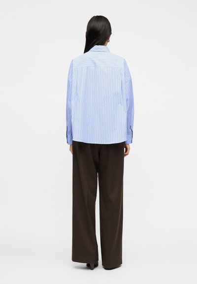 Γυναίκα με μακριά σκούρα μαλλιά που φοράει μπλε πουκάμισο με λεπτές ρίγες και φαρδιά καφέ παντελόνια, στέκεται με την πλάτη προς τα έξω μπροστά σε απλό φόντο.