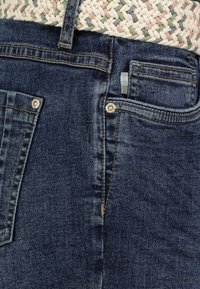 Die Jeans aus Denim haben eine dunkelblaue Farbe, mit sichtbaren Nähten und silbernem Zubehör. Sie verfügen über Gesäßtaschen und einen gewebten Gürtel in natürlichen Farbtönen.