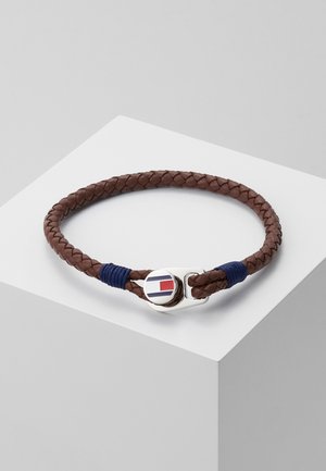 Hnědý pletený kožený náramek se stříbrnou sponou, na které je kulatý emblém se pruhy v červené, bílé a modré barvě, ležící na bílém podstavci.