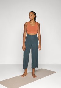 Oransje crop top og mørk teal vide bukser laget av myk stoff, vist på en barføtt figur som står på en yogamatte.