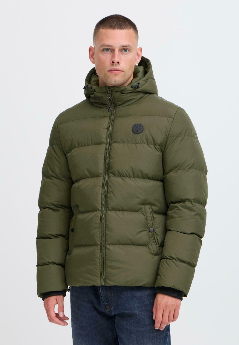 Man wearing olive green quilted puffer jacket with hood, standing against a plain light background.