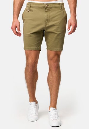 Mand iført olivengrønne knælange shorts med knapper foran og hvide sneakers, stående mod en ensfarvet baggrund.