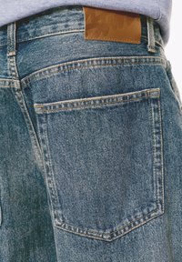 Primo piano di una tasca di jeans in denim blu e di una toppa in pelle marrone sul cinturino, indossati con una maglietta di un leggero colore viola.