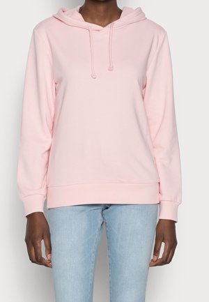 Person iført en lyserød hættetrøje med snore og lyseblå jeans, stående foran en ensfarvet baggrund.