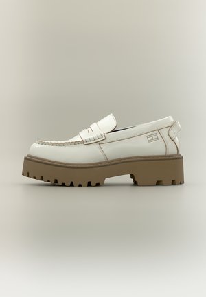 Mocassin en cuir blanc avec semelle épaisse à crampons marron, coutures apparentes et languette de traction sur un fond neutre.