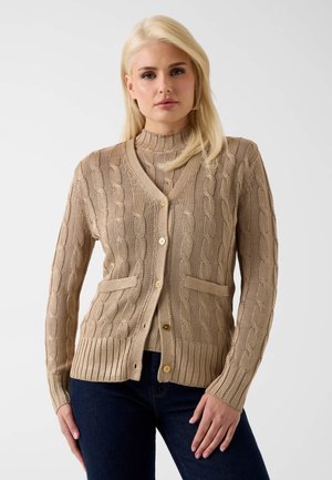 Blondynka ubrana w beżowy sweter na guziki z warkoczowym wzorem, nałożony na pasujący golf, do tego ciemnoniebieskie dżinsy.